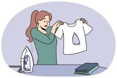 Beceriksiz ev hanımı tişörtündeki yanık deliği ütülüyor ve hatayı nasıl düzelteceğini bilmiyor. Kadın ütü masasının yanında duruyor. Ütü ve ütüyle. Hasarlı eşyalara bakıyor..