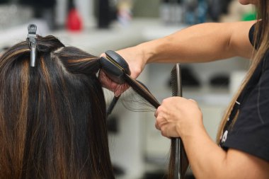 Roma, İtalya - 10 31 2024 Genç bir kuaför, genç bir kadının saçını bir kadın kuaföründe düzleştirmek için ütü kullanıyor. 
