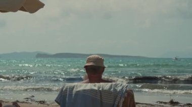 Korfu adası, Yunanistan - 2023.07.01 - 09: Güzel bir sahilde güneşli bir yatakta güneşlenen yaşlı bir adam, sıcak, güneşli gökyüzünün altında, mavi su arka planında hafif dalgalarla