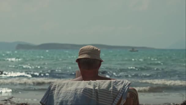 希腊科孚岛 2023 老人在美丽的海滩上 在炎热 阳光灿烂的蓝天下 在蓝色水底的柔和波浪中 欣赏着一本关于日光浴床的书 — 图库视频影像