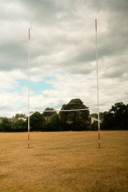 Chiswick, Londra, İngiltere 2025.08.05 Kuru, sararmış bir yaz sahasının üzerinde duran uzun rugby kaleleri dramatik, bulutlu bir gökyüzünün altında arka planda evler ve ağaçlar var..