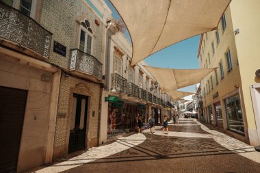 Algarve, Portekiz 2025.06.28 Pembe tişörtlü yaşlı bir adam Algarves 'in göbeğindeki mozaik kaplı bir alışveriş sokağının gölgesinde oturuyor.             