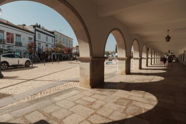 Tavira, Algarve, Portekiz 2025.07.02: Kemerli sütunlar, sallanan bayraklar ve güneşli ana meydanda çağdaş bir taş çeşme ile beyazlatılmış belediye binası                                                   