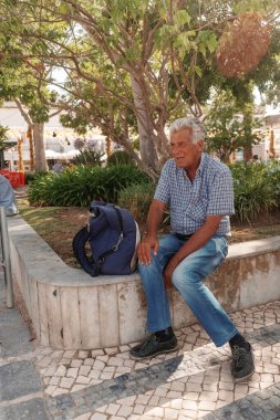 Algarve, Portekiz 2025.06.28 Pembe tişörtlü yaşlı bir adam Algarves 'in göbeğindeki mozaik kaplı bir alışveriş sokağının gölgesinde oturuyor.             