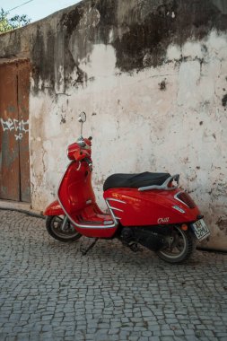 Tavira, Algarve, Portekiz 2025.07.02: Parlak kırmızı scooter taşlı bir caddede soluk boya ve grafiti ile yıpranmış bir duvarın yanında park edilmiş.. 