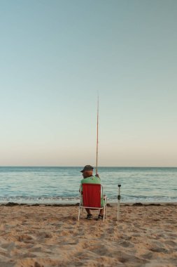 Algarve, Portekiz 2025.07.03: Bir adam elinde oltayla bir sandalyede oturuyor, berrak bir akşam gökyüzünün altında sakin bir denize bakıyor, ay ufuktan yükselirken.. 