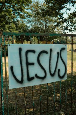 Chiswick, Londra, İngiltere - 2025.08.12 - Paslı metal bir kapıda sprey boyalı İsa tabelası, yaz güneşinde park ve yeşil ağaçlar sergilendi.