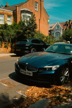 Chiswick, Londra, İngiltere - 2025.08.12 - Arka planda siyah bir Porsche SUV ve tuğla evler ile güneşli bir iskan caddesinde kaldırım kenarına park edilmiş mavi BMW Z4.