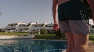 Algarve, Portekiz 2025.06.25 Yüzme şortlu bir adam güneşli bir otelde açık havuz kenarında duruyor, beyaz apartman binaları ve arka planda manzaralı bahçeler var.. 
