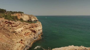 Algarve, Portekiz 2025.06.25 dramatik altın kayalıklar turkuaz Atlantik sularıyla buluşuyor ve ufukta berrak bir yaz gökyüzü altında beyaz bir yelkenli var.. 