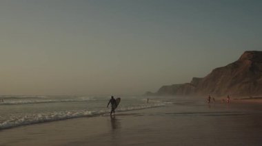 Algarve, Portekiz 2025.06.25 Sörfçü elinde kayalıkların üzerinde altın gün batımı ışığı ve nazik dalgalarla bir tahtayla sudan çıkıyor.. 