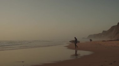 Algarve, Portekiz 2025.06.25 Dalgıç kıyafetli sörfçü altın saat boyunca kumlu kıyı şeridi boyunca yumuşak dalgaları ve engebeli kayalıkları olan bir tahta taşıyor.. 