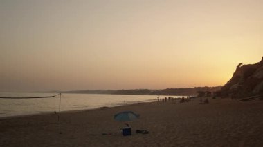 Algarve, Portekiz 2025.07.01 Praia da Falsia üzerinde gün batımı, voleybol ağı, şemsiye ve ikonik kırmızı kayalıkların yakınındaki plajın tadını çıkaran insanlar.. 