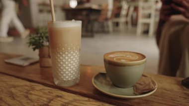 Sagres, Algarve, Portekiz 2025.07.01 Bir buzlu latte 'nin yakın planında bir şablonlu bardakta ve bir kapuçino ile sade ahşap bir masa üzerinde latte sanatı. 