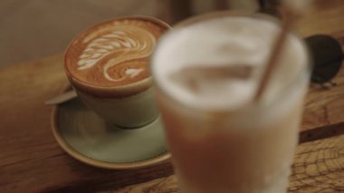 Sagres, Algarve, Portekiz 2025.07.01 Bir kapuçino Artistik shot odaklı latte sanat, uzun buzlu latte ön planda bulanık bir ahşap kafe masa üzerinde. 