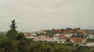 Silves, Algarve, Portekiz 2025.07.01 Silves kasabasının beyaz evleri, kilisesi ve bulutlu gökyüzünün altındaki tarihi kırmızı kumtaşı kalesi.. 