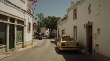 Estoi, Algarve, Portekiz 2025.07.01 Sarı bir BMW çatıda çizgili bir yastık ile dar bir cadde boyunca güneşli bir günde yıkanmış binalarla kaplı..