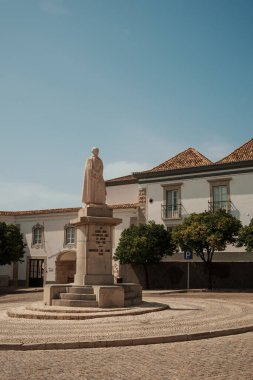 Algarve, Portekiz 2025.06.28 Güneşli bir günde tarihi binalar ve portakal ağaçlarıyla çevrili taş bir meydandaki taş heykel. 