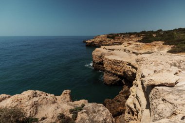 Algarve, Portekiz 2025.06.29 Engebeli kireçtaşı kayalıkları ve açık bir yaz gökyüzü altında derin mavi Atlantik sularının panoramik manzarası. 