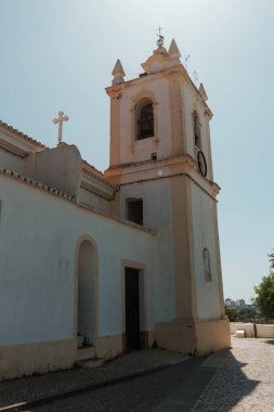 Algarve, Portekiz 2025.06.30 Çan kulesi ve perende dini heykeli olan tarihi kilise, öğlen güneşinin altında kaldırım taşı meydanında yakalandı.. 
