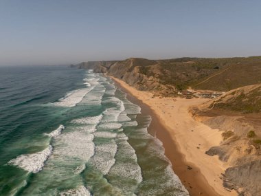 Algarve, Portekiz 2025.07.01 Praia da Amoreira insansız hava aracı görüntüsü. Turkuaz dalgalar engebeli kayalıklar ve doğal kıyı manzarasıyla desteklenen geniş kumlu bir plaja doğru ilerliyor.. 
