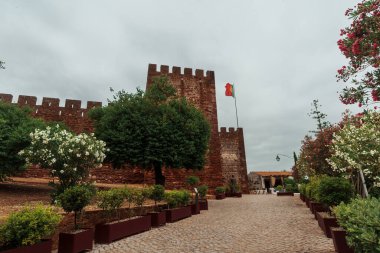 Silves, Algarve, Portekiz 2025.07.04 Silves Kalesi manzaralı kırmızı kumtaşı duvarlar Portekiz bayrağı, kaldırım taşı yolu ve çiçek açan ağaçlar ön planda.. 
