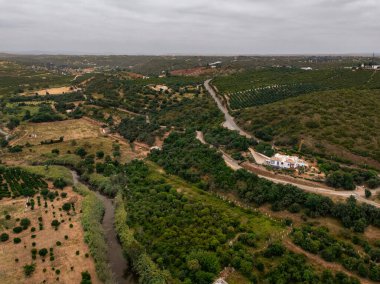 Silves, Algarve, Portekiz 2025.07.04 Sarmal nehir, yeşil tarlalar, meyve bahçeleri, tepeler ve Silves yakınlarındaki bir kır evi ile kırsal arazi görüntüsü 
