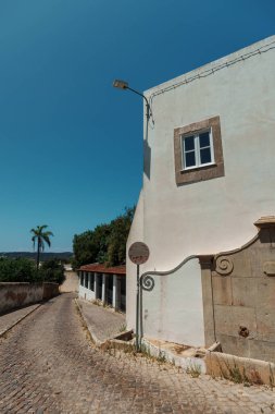 Silves, Algarve, Portekiz 2025.07.05 Temiz bir yaz gökyüzünün altında tarihi fıskiye duvarı, taş detayları ve palmiye ağacı olan sessiz bir cadde.. 
