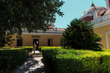 Faro, Algarve, Portekiz 2025.07.05 Taş heykel çeşmesi, budanmış çitler ve kubbeli sarı tarihi bina manzaralı bahçe.. 
