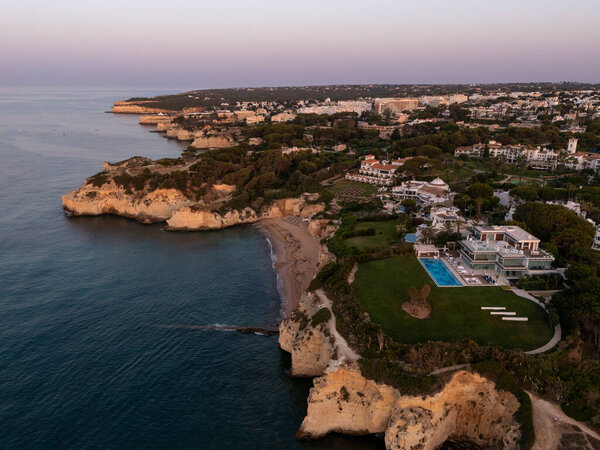 Алгарве, Португалия 2025.07.01: Вид на беспилотник на рассвете роскошных вилл с бассейнами на скале с видом на уединенный песчаный пляж и золотые скалы вдоль Атлантического побережья. 