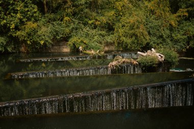 London, UK  2025.08.09  A stepped waterfall cascades through multiple concrete tiers, surrounded by lush summer trees and floating driftwood along the canal 