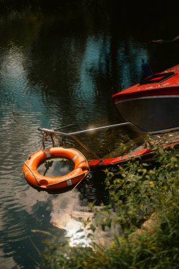 London, UK  2025.08.09  A close-up view of an orange lifebuoy floating on calm canal waters beside a partially sunken red boat, reflecting sunlight and greenery. 