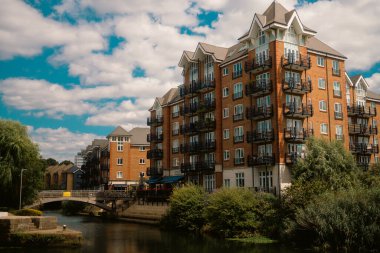 London, UK  2025.08.09  Red brick apartment buildings with black balconies stand along the canal beside a pedestrian bridge under a partly cloudy summer sky. 
