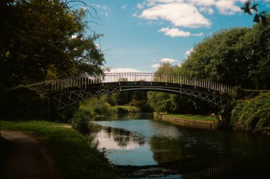 Londra, Birleşik Krallık 2025.08.09 Tarihi bir demir köprü sulara yansıyan bulutlarla dolu yemyeşil ve mavi gökyüzü tarafından çerçevelenmiş sakin bir kanal boyunca uzanır. 