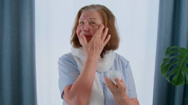 Yaşlı kadın yüz nemlendiricisini yüz derisine sürüyor, yaşlanmayı önleme süreci duruyor. Kırışıklıklara karşı özel muamele gören yaşlı bir kadın.