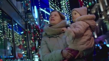 Noel arifesinde annesiyle yürüyen tatlı bir kız bebek, Noel süsü ve sokakta ışıklar. Anne ve bebekle yeni yıl akşamı