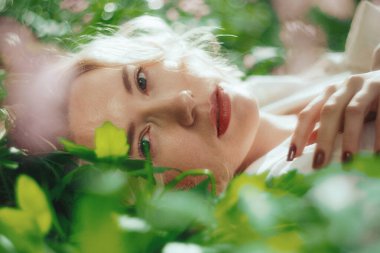 Ormanda yeşil çimlerin üzerinde yatan güzel sarışın kadın, parfüm reklamı konsepti. Romantik bir havada gülümseyen genç kız.
