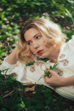 Ormanda yeşil çimlerin üzerinde yatan güzel sarışın kadın, parfüm reklamı konsepti. Romantik bir havada gülümseyen genç kız.