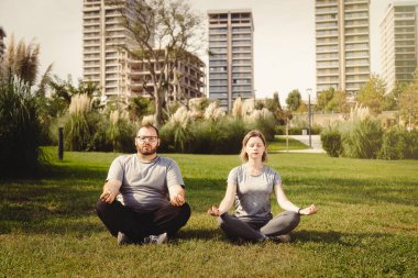 Park lotusu pozisyonunda meditasyon yapan iki kişi, aşırı kilolu bir adamla çim beyaz kadın eğitmen üzerinde sabah sporu antrenmanı yapıyor.