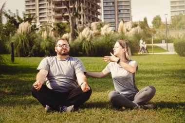 Park lotusu pozisyonunda meditasyon yapan iki kişi, aşırı kilolu bir adamla çim beyaz kadın eğitmen üzerinde sabah sporu antrenmanı yapıyor.