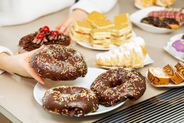 Kadınların eli, tatlı yiyeceklerle çikolatalı donutları masadan alıyor. Aşırı yeme sorunları ve şeker bağımlılığı