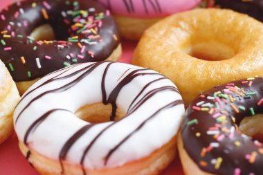 Çeşit çeşit çikolatalı donutlar, üzerine sos serpilmiş renkli çörekler ve çok renkli tatlı çöreklerin karışımı pembe arka planda serpiştirilmiş. en üstteki görünüm