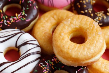 Çeşit çeşit çikolatalı donutlar, üzerine sos serpilmiş renkli çörekler ve çok renkli tatlı çöreklerin karışımı pembe arka planda serpiştirilmiş. en üstteki görünüm