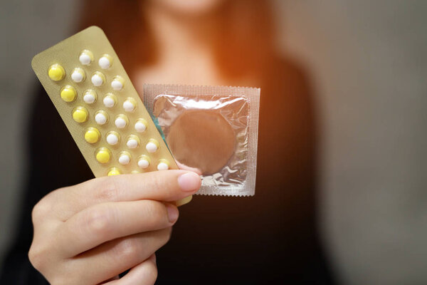 Contraceptive means: Woman hand holding contraceptive pills and condom sitting on sofa background. Protection, safe sex. Contraception, concept birth control. Copy space