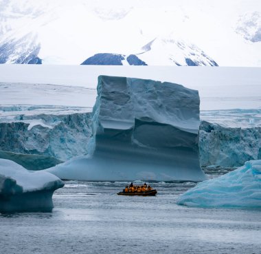 Antarktika seferi, sarı parkalı seyahat yolcuları dev beyaz bir buzdağına çok yakın şişme bir Zodiac botla seyahat ediyorlar. İklim değişikliği ve küresel ısınma