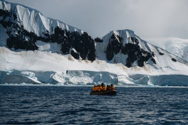 Antarktika seferi, sarı parkalı seyahat yolcuları dev beyaz bir buzdağına çok yakın şişme bir Zodiac botla seyahat ediyorlar. İklim değişikliği ve küresel ısınma