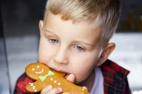 Cute caucasian kid eats gingerbread and makes a funny face expression. Christmas food. Atmospheric, aesthetic Christmas home for New Year's Eve. Cheerful baby boy. Joyful, good mood.