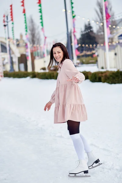 Happy smiling stylish brunette in dress ice skating outdoor in winter. Festive holidays mood. Dreams come true concept.
