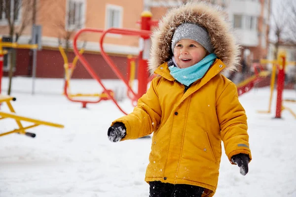 Happy child with snowy face having fun in the snow in the city. Winter fun outside. Boy in bright orange winter Jacket. High quality photo