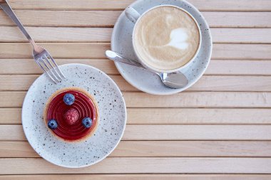 French fresh sweet blueberry and raspberry tart on white ceramic plate with cup of cappuccino. Coffee time, breakfast. Copy space on wooden background